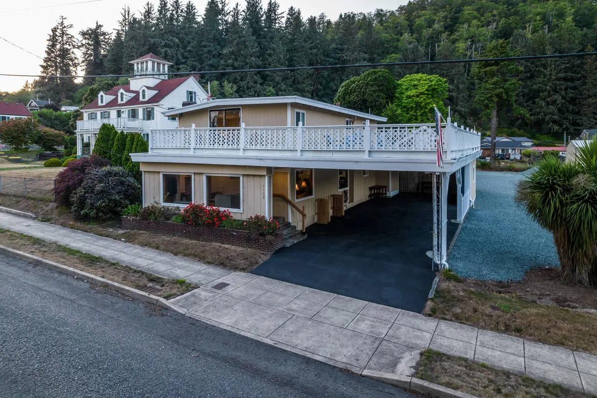 A wide view of the property shows the wraparound balcony, a landscaped garden, a classic vintage-style house, and a driveway with several parked vehicles. The composition highlights the interconnectedness of outdoor spaces, from arrival to relaxation on the upper deck.