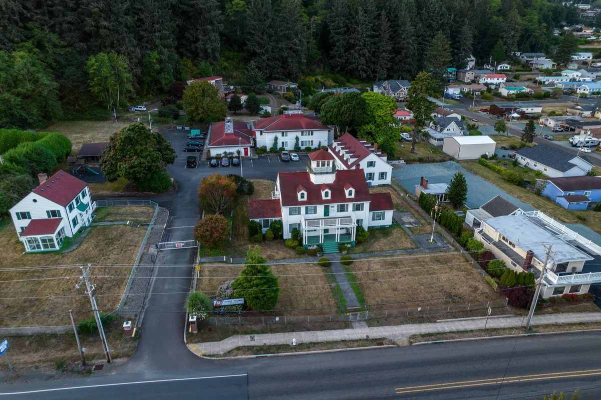 The image shows a historic white house with red roofs, nestled among greenery and featuring a clearly visible parking area. The open outdoor space suggests ample room for vehicles, boats, or RVs, highlighting the convenience for guests with outdoor gear.