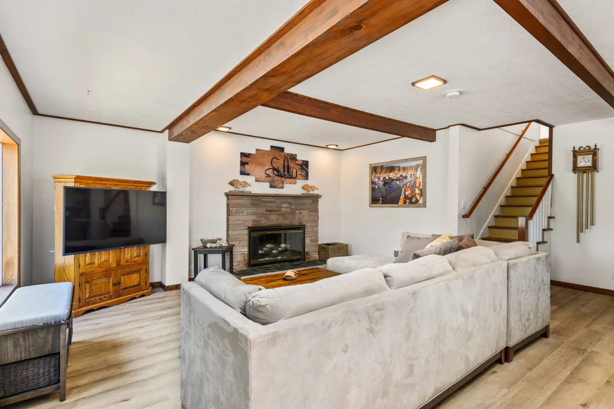 The image showcases a warm, inviting living room featuring a crackling fireplace as the centerpiece, a plush sectional sofa, a mounted television, tasteful coastal artwork, and a staircase leading to the upper floor. The space is designed for relaxation and gathering, making it ideal for families or groups to unwind together after a day of coastal adventures.