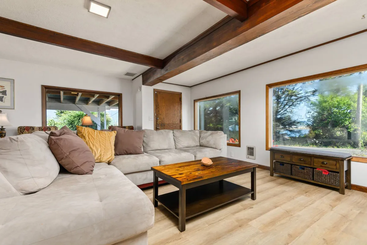 A cozy living room with a plush sofa, wooden coffee table, large windows letting in natural light, and exposed wooden beams overhead. The space exudes a welcoming, beachy vibe—sunlight floods the room, and comfortable seating invites conversation or relaxation.