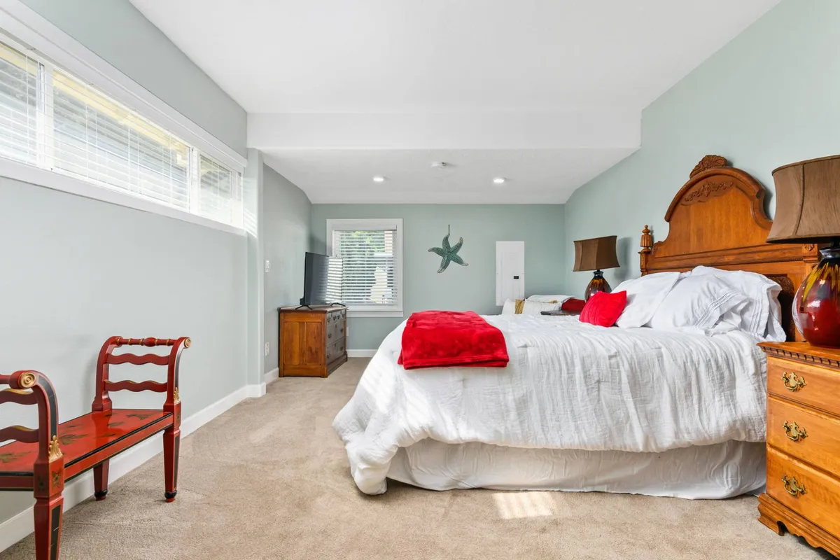 A restful bedroom featuring a plush bed with white linens and vibrant red accents. The room includes wooden nightstands, a red and black chair, and sunlight streaming through the windows, creating a calm and cozy atmosphere ideal for unwinding after a long day.