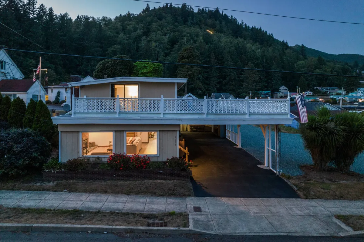 An inviting exterior view of a house with a balcony overlooking a landscaped front yard. The illuminated windows suggest warmth and welcome, while the balcony offers a perfect vantage point for group gatherings and sunset views.