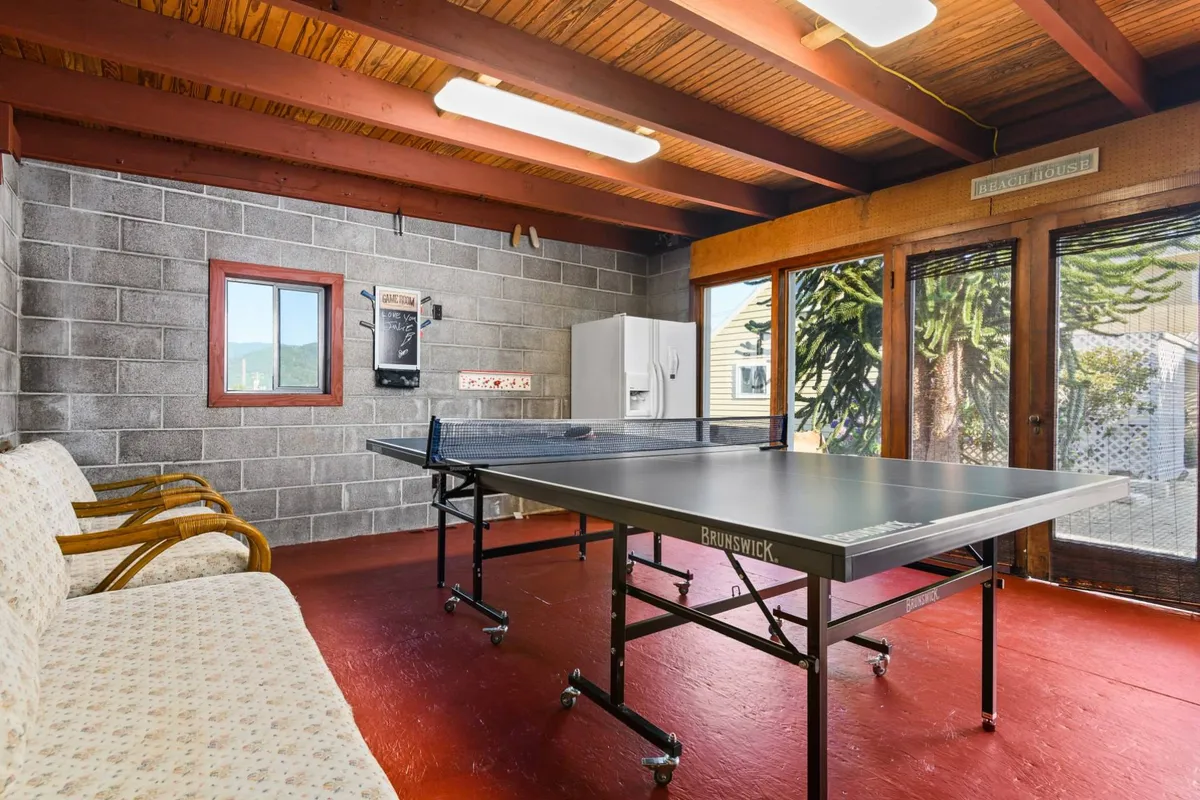 This interior image features a lively game room with a central ping pong table, a comfortable couch set against large windows that let in natural light, and a casual, playful setup ideal for socializing or family fun. The layout encourages group activities and underscores the recreational focus of the space.