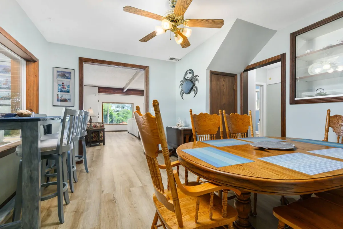 A dining space with a large wooden table, bar stools at the kitchen counter, coastal-themed decor (including a decorative crab on the wall), and expansive windows that flood the area with sunlight. The layout invites conversation and togetherness.