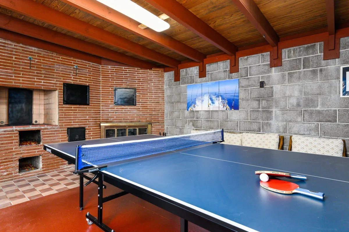 A cozy game room interior with a ping pong table as the centerpiece, complemented by a brick fireplace and decorative wall art. The room exudes a playful yet relaxed mood, ideal for both spirited games and winding down after a busy day.