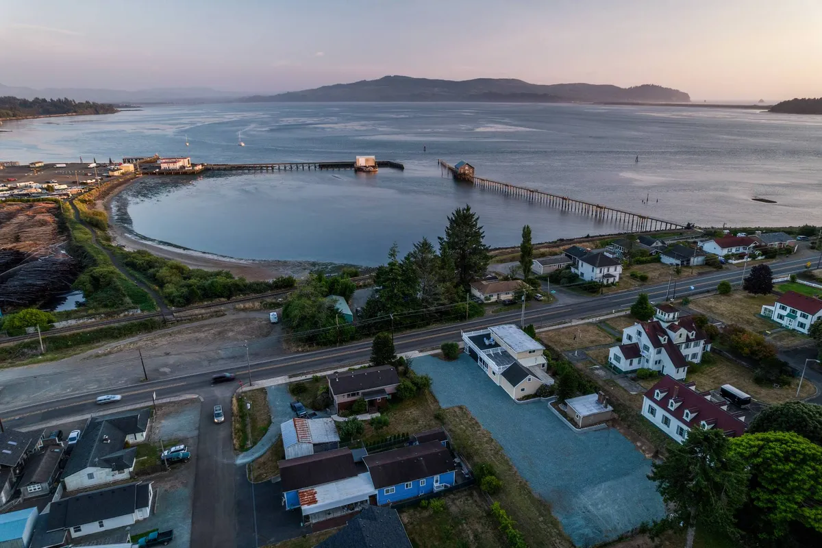 This image captures the scenic pier, circular bay, nearby houses, and the road leading to the water. The tranquil setting emphasizes easy access to the waterfront for boating or fishing, with visible community activity along the shore, and surrounding natural beauty.