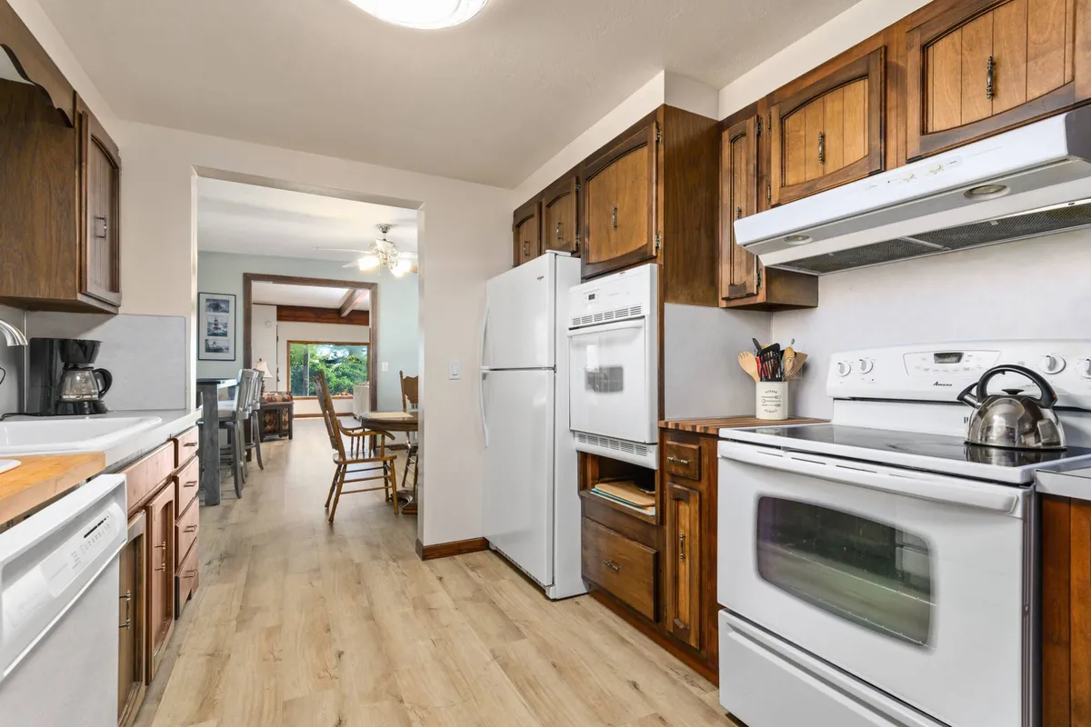 A view from the kitchen featuring modern appliances, warm wooden cabinetry, and a glimpse of the dining area beyond. The scene suggests a hub for cooking, sharing meals, and gathering with family or friends in a bright, welcoming environment.