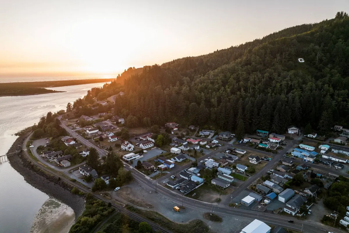 A picturesque sunset view over water, with small town houses nestled against forested hills and a river winding through the landscape. The scene evokes the natural beauty and serenity of the Oregon Coast, ideal for group outings, exploration, and capturing memorable moments.