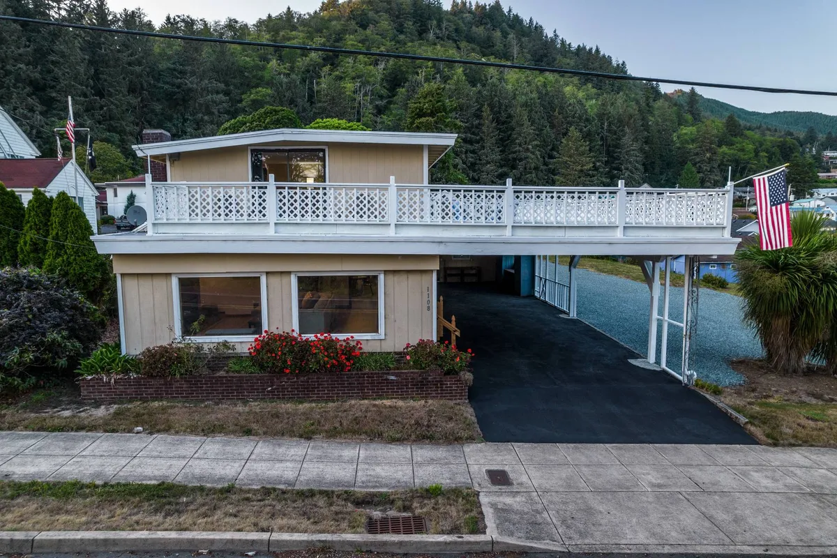 This exterior photo captures the balcony with a white railing adorned with the American flag, a beautifully landscaped garden with vibrant flowers, and a spacious parking area in the foreground. The setting is bright and inviting, emphasizing both the home's welcoming entrance and its practical outdoor amenities.